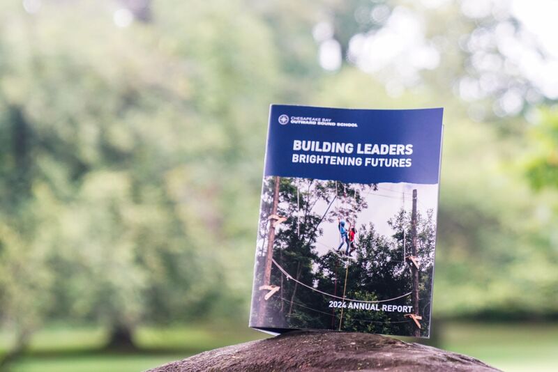 The image shows a book or report titled "BUILDING LEADERS BRIGHTENING FUTURES" and "2026 ANNUAL REPORT". The cover has a picture of people on a ropes course, set against a backdrop of trees and greenery. The book is placed on a rock or stone surface, with a blurred background of more trees and foliage, suggesting an outdoor setting.